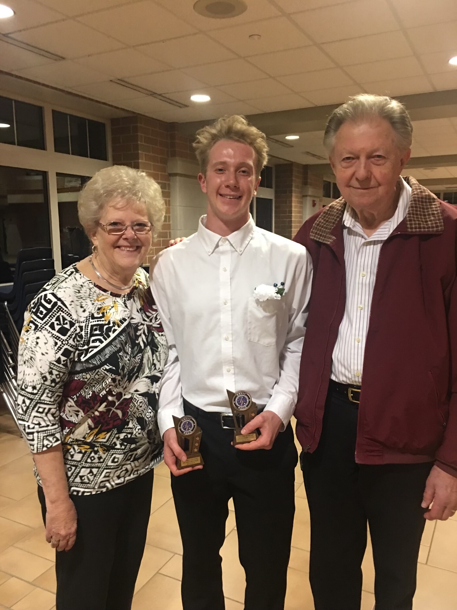 Three people smile for a photo at an awards event: a young man in a white shirt holding trophies, flanked by an older woman and man in a burgundy jacket.