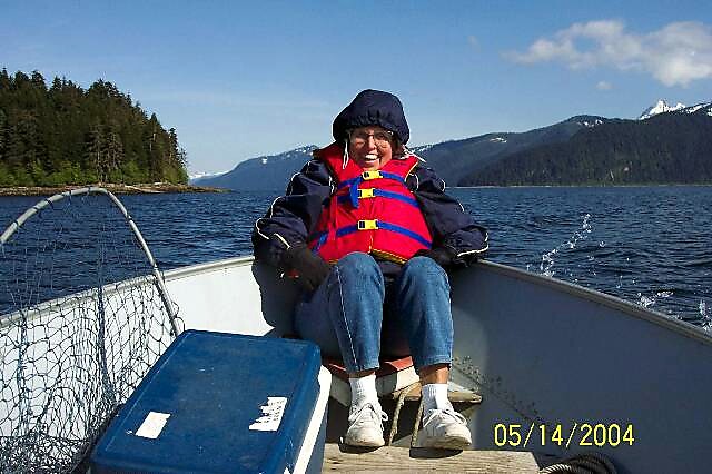 Woman wearing a red life vest sits at the bow of a small boat on a lake with forested hills and mountains in the background; date 05/14/2004.
