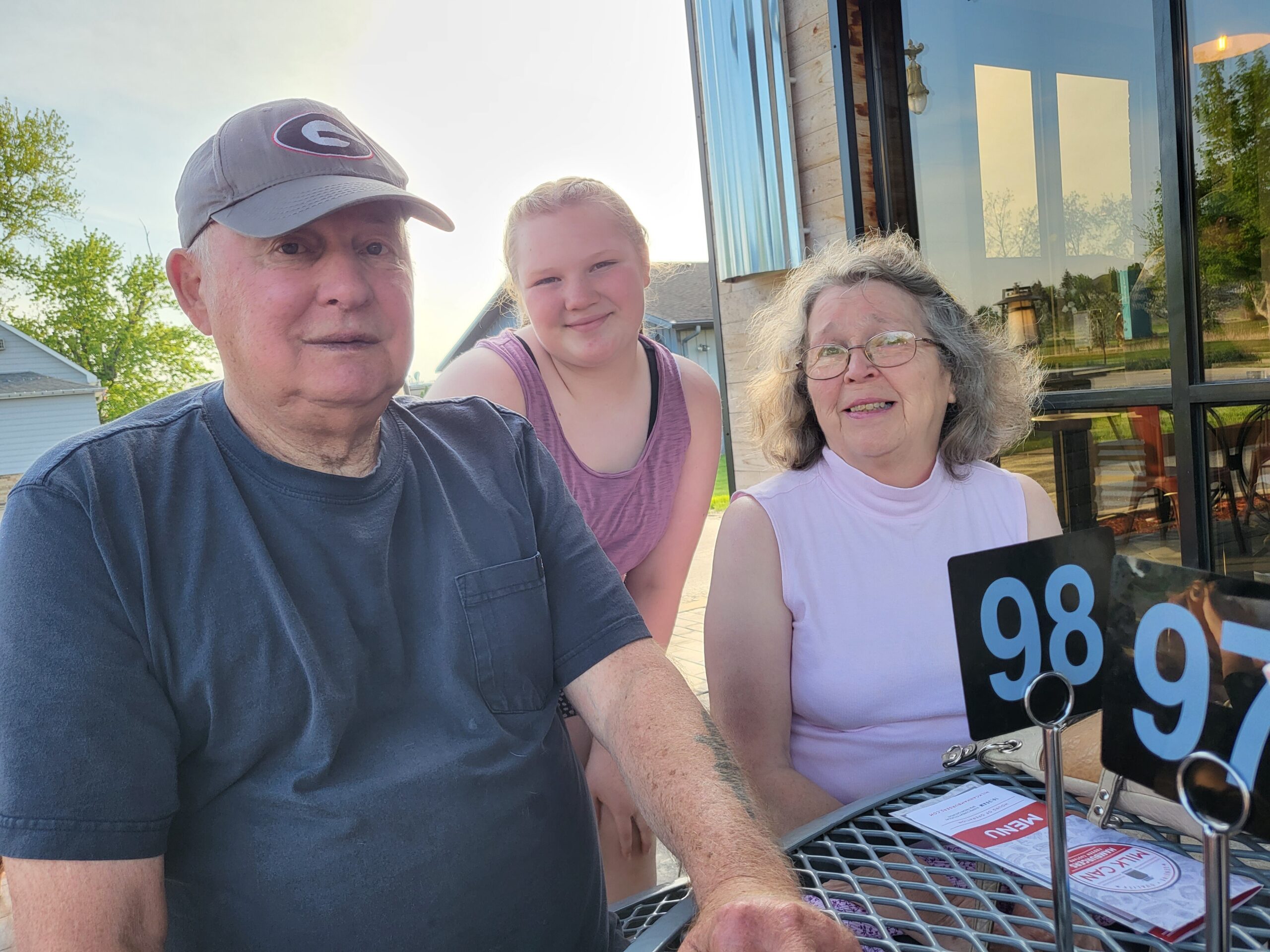 Three people, an older man, a young girl, and an older woman, pose for a selfie outside a building with glass windows, smiling at the camera.