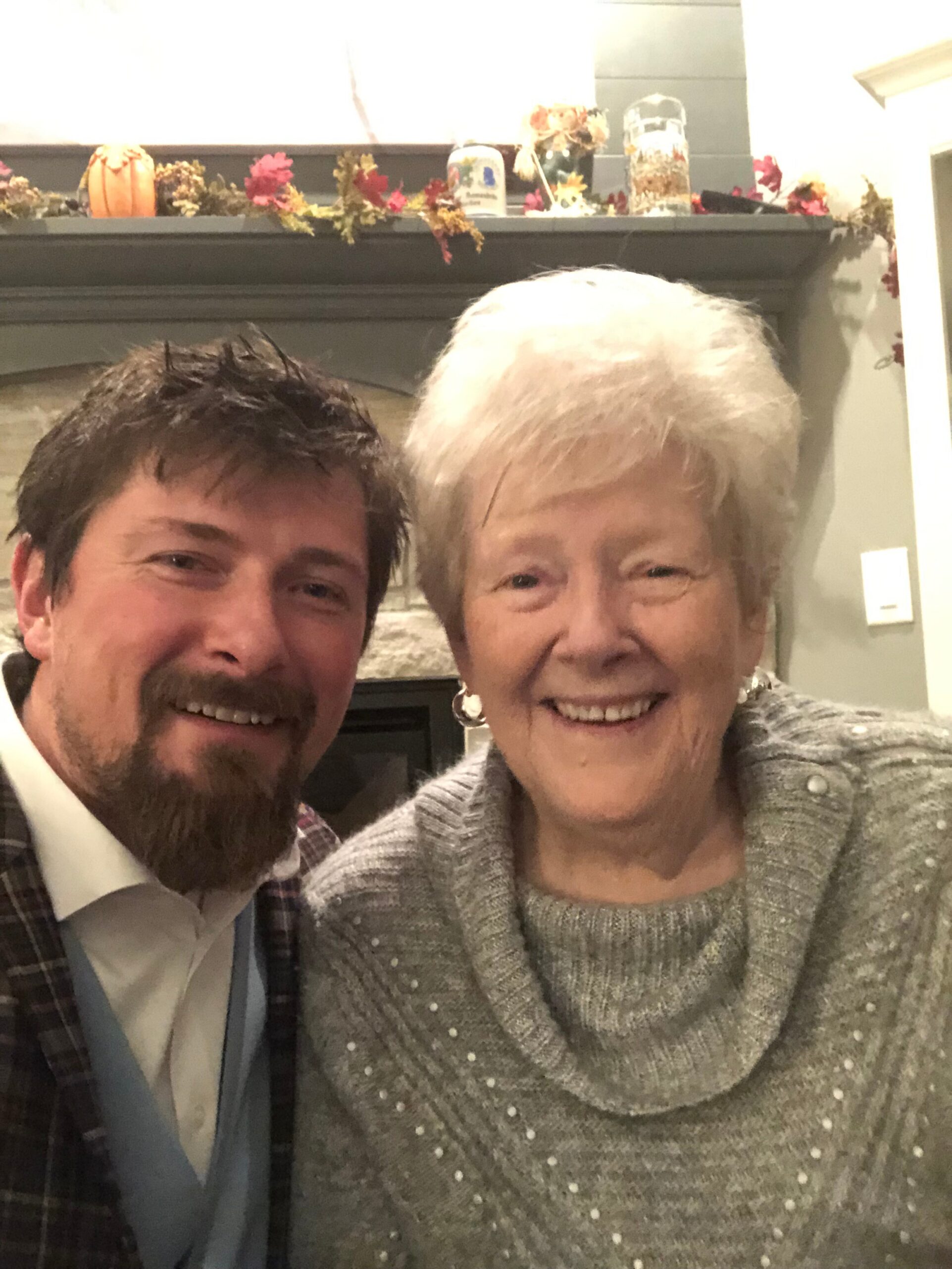 Two people smiling for a selfie in a living room with autumn pumpkin and garland decorations on the mantel behind them.