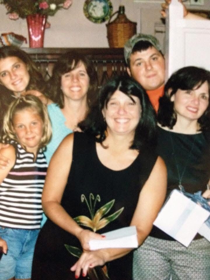 Group of smiling people posed closely for a photo indoors at a gathering, with a woman in a black dress holding a white box or gift.