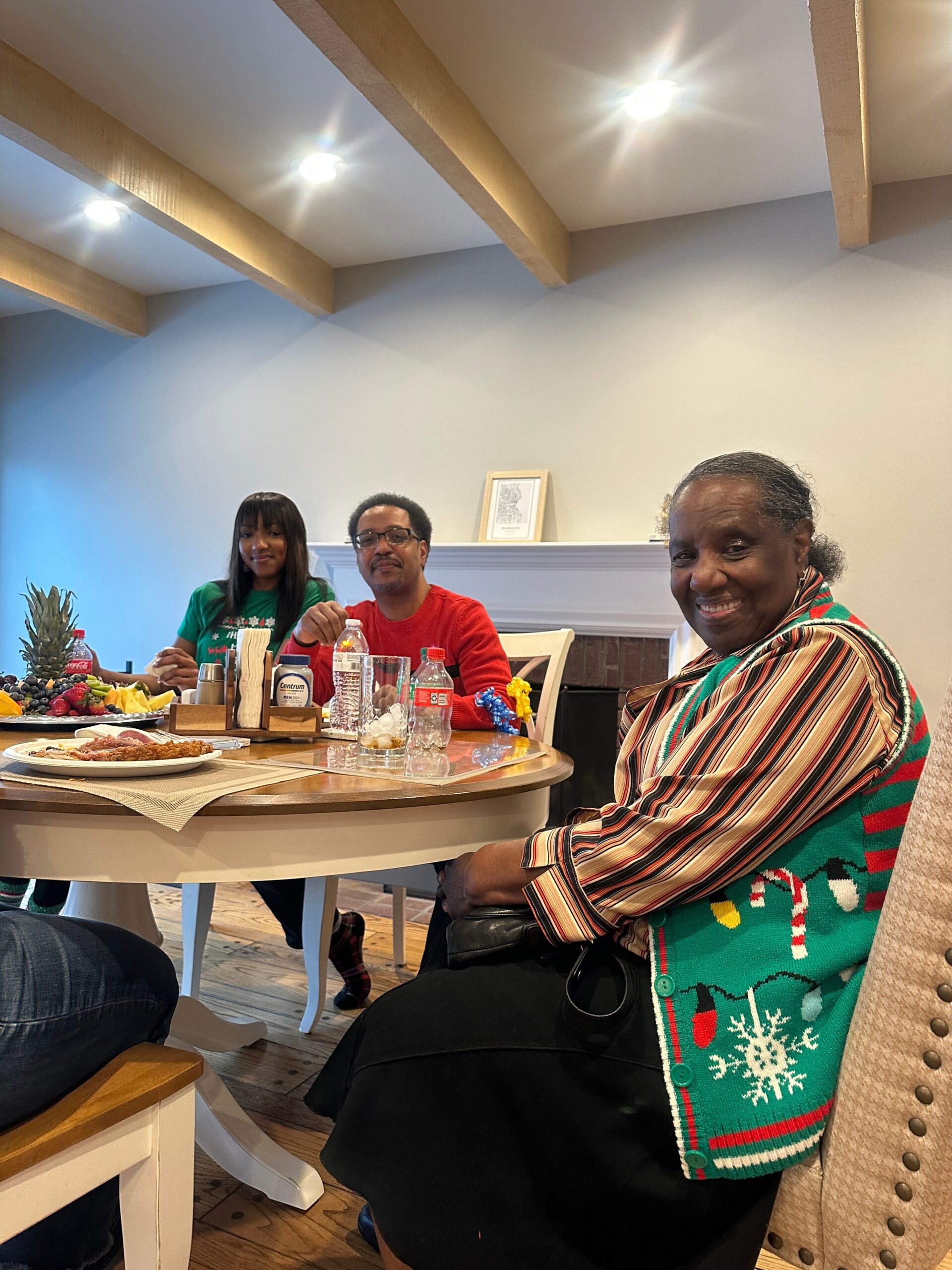 Three people sit around a dining table with fruit, drinks, and foods, smiling indoors near a warm hearth. The room has exposed wooden ceiling beams.