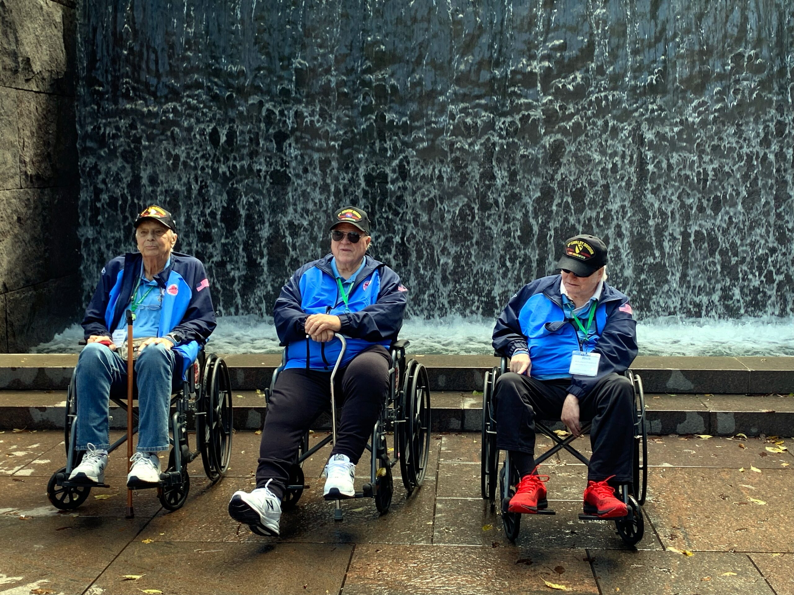 Three elderly men in wheelchairs sit in a row in front of a large cascading water feature outdoors, wearing blue jackets and caps.