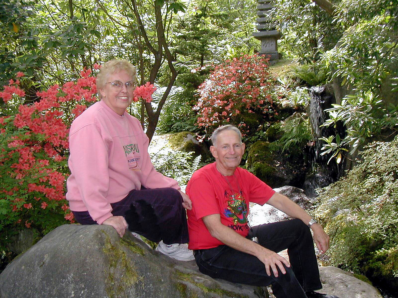 Two adults sit on a large rock in a lush garden, with pink azalea blooms and a small waterfall in the background.