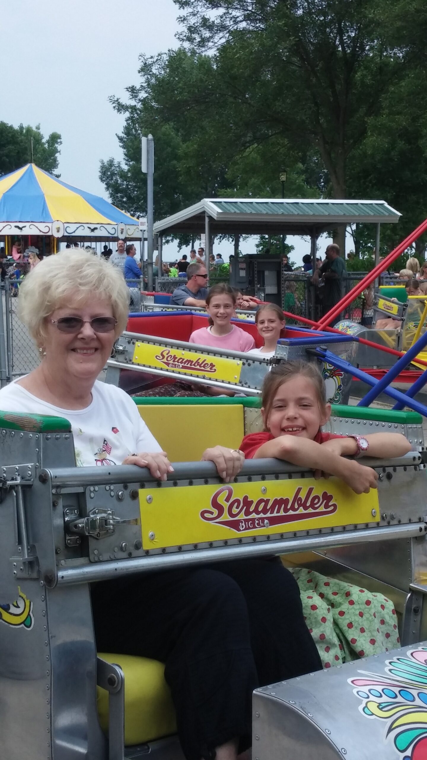 Smiling older woman with short light hair and two girls sit in a Scrambler amusement ride car at a crowded fairground.