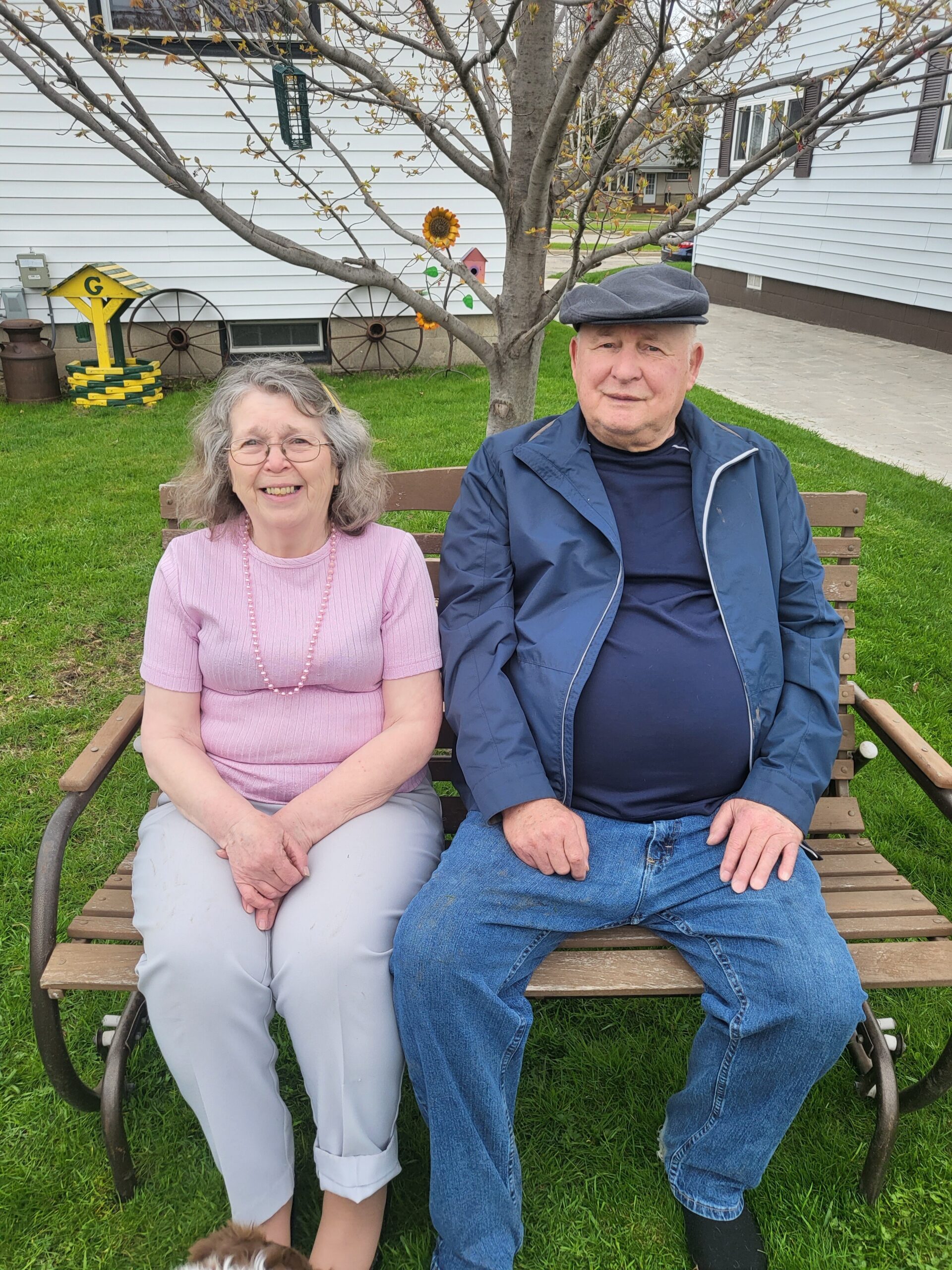 Smiling elderly couple sits on a wooden bench in a front yard with a budding tree and a yellow birdhouse in the background.