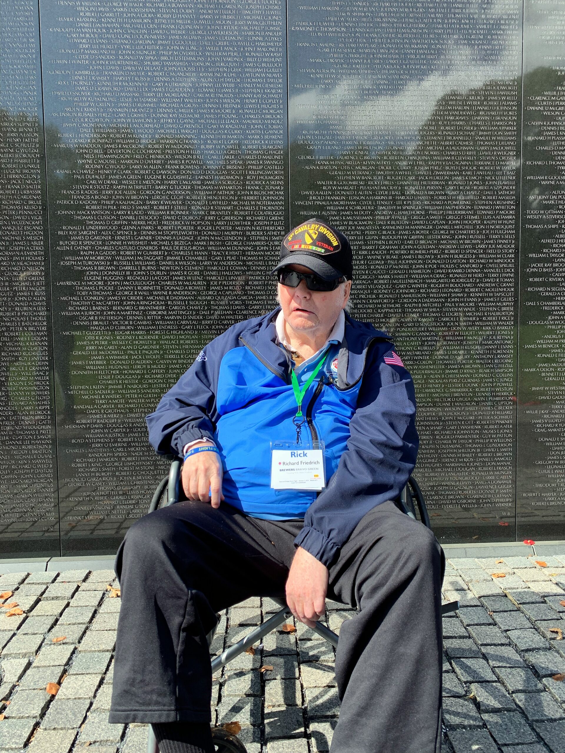 Older man in a wheelchair wearing a blue jacket and sunglasses, in front of a wall engraved with numerous names.