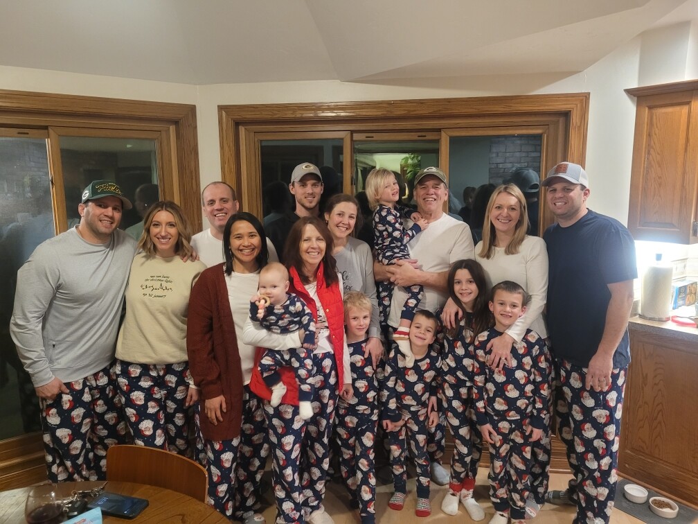 Group of adults and children in matching Christmas pajama pants posing in a home kitchen for a holiday gathering.