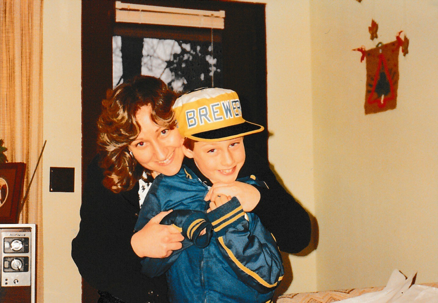 Mother hugging a smiling boy in a blue jacket indoors; he wears a yellow cap with 'BREWER' on it.
