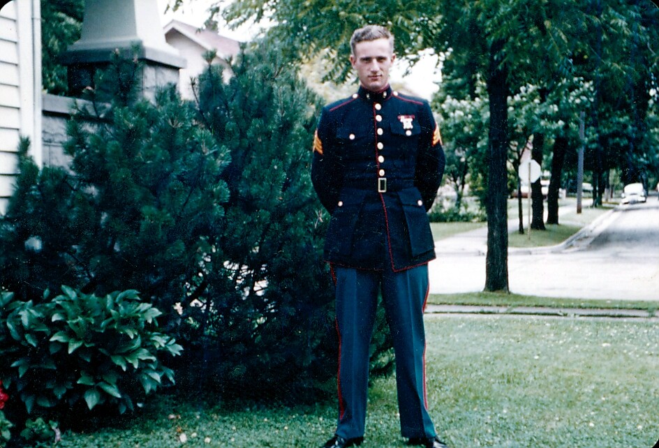 A man in dress military uniform (dark jacket with gold buttons and medals) standing on a lawn in a suburban neighborhood.