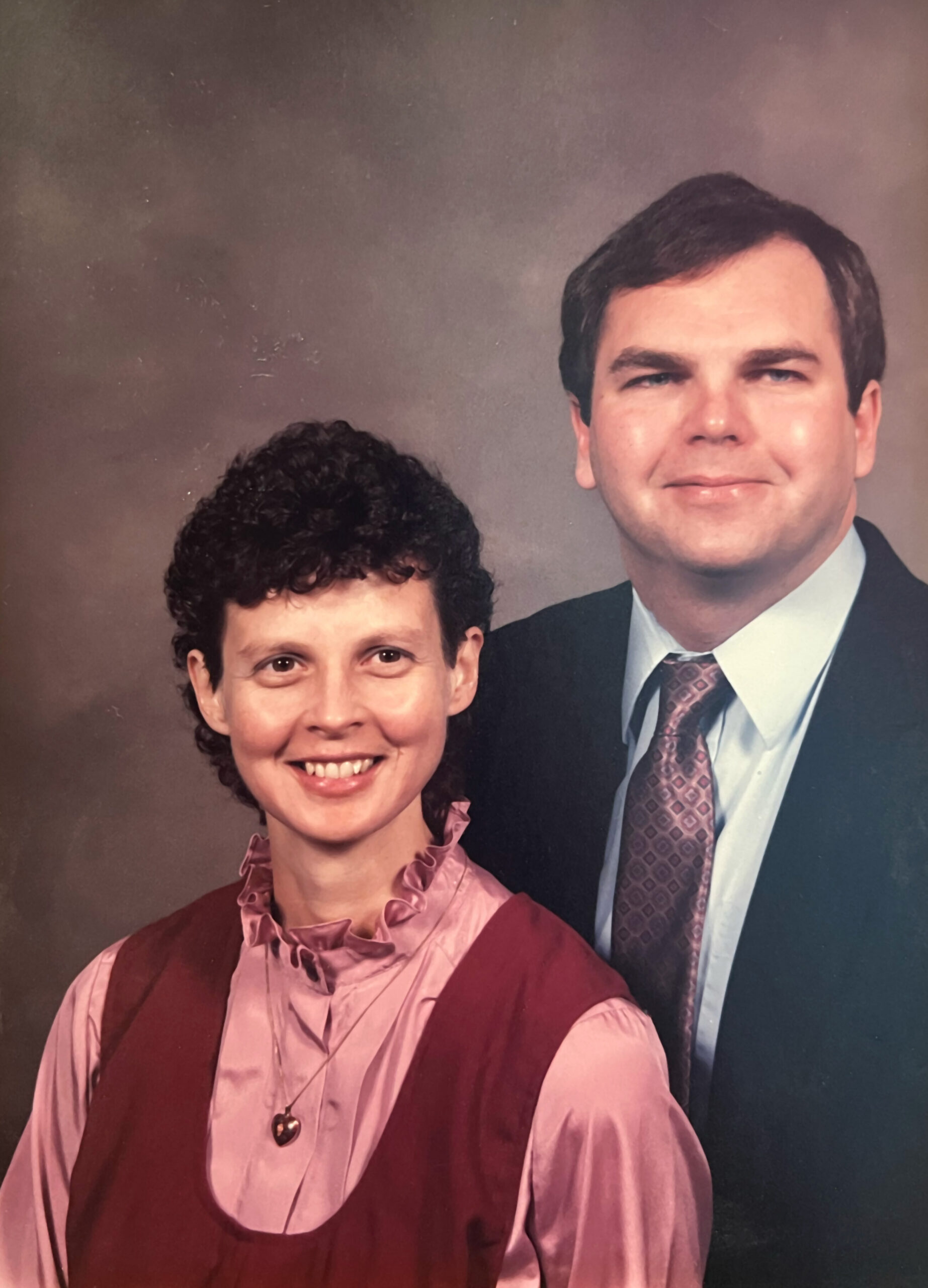 Smiling woman in pink blouse with burgundy vest and heart pendant poses with a man in a suit and patterned tie in a studio portrait backdrop.