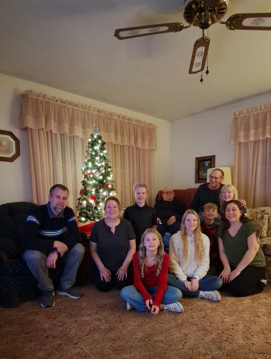 Group of eleven people, adults and children, posing together in a living room around a lit Christmas tree.
