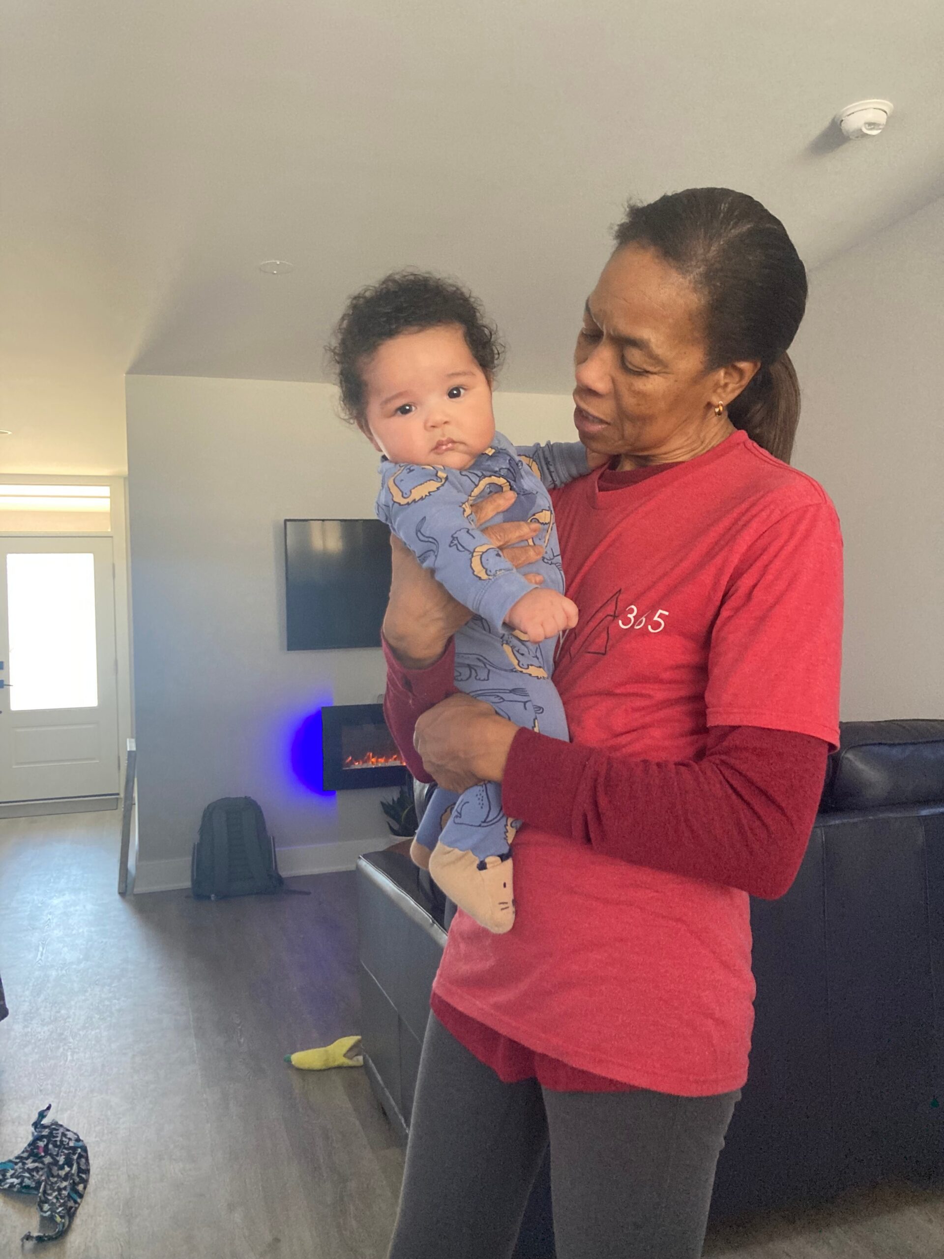 Woman in red shirt holding a baby in a living room, facing left with a sofa and TV behind them.