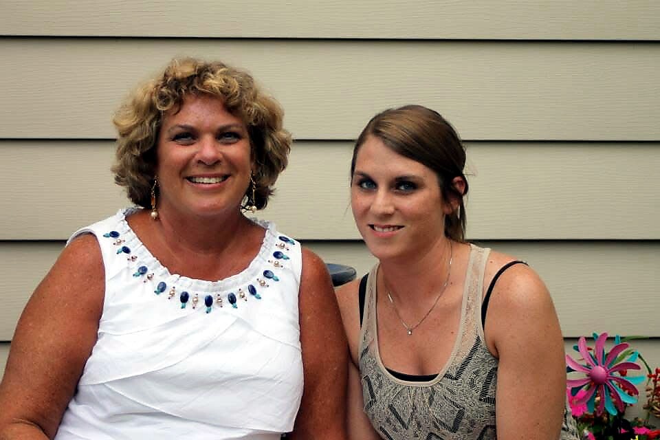 Two women smiling and posing outdoors against a beige house; older woman in a white sleeveless top with blue bead embellishments, younger woman in a gray tank top; a colorful pinwheel in view nearby.