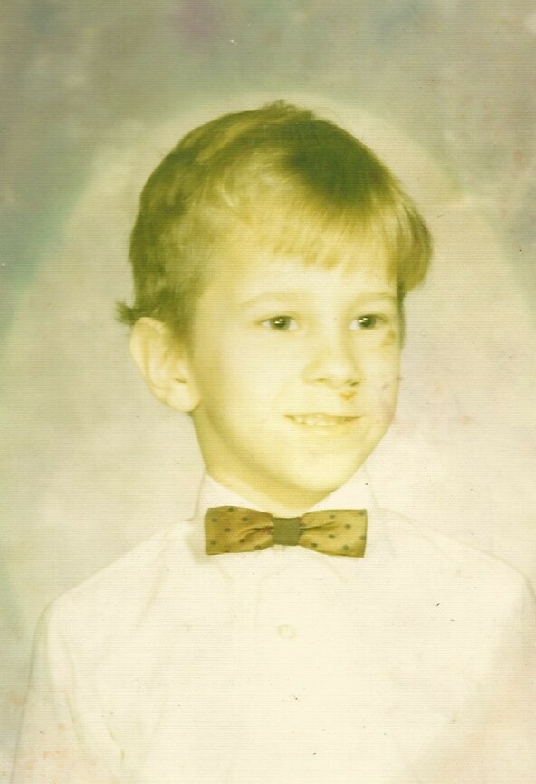 Young boy in a white shirt with a polka-dot bow tie, smiling for a formal portrait.