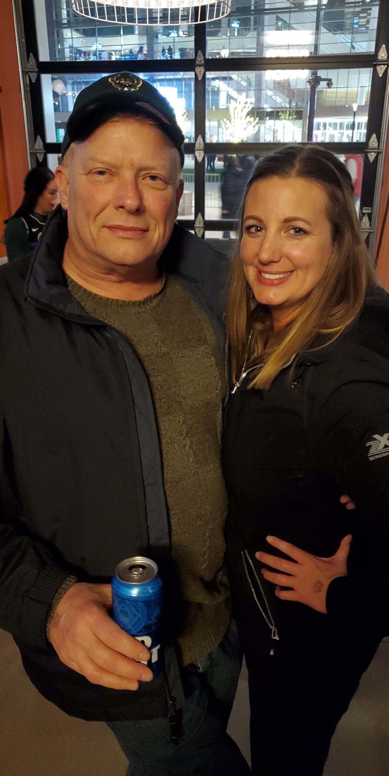 Two adults posing for a selfie indoors, man holding a blue beverage can and woman smiling beside him.