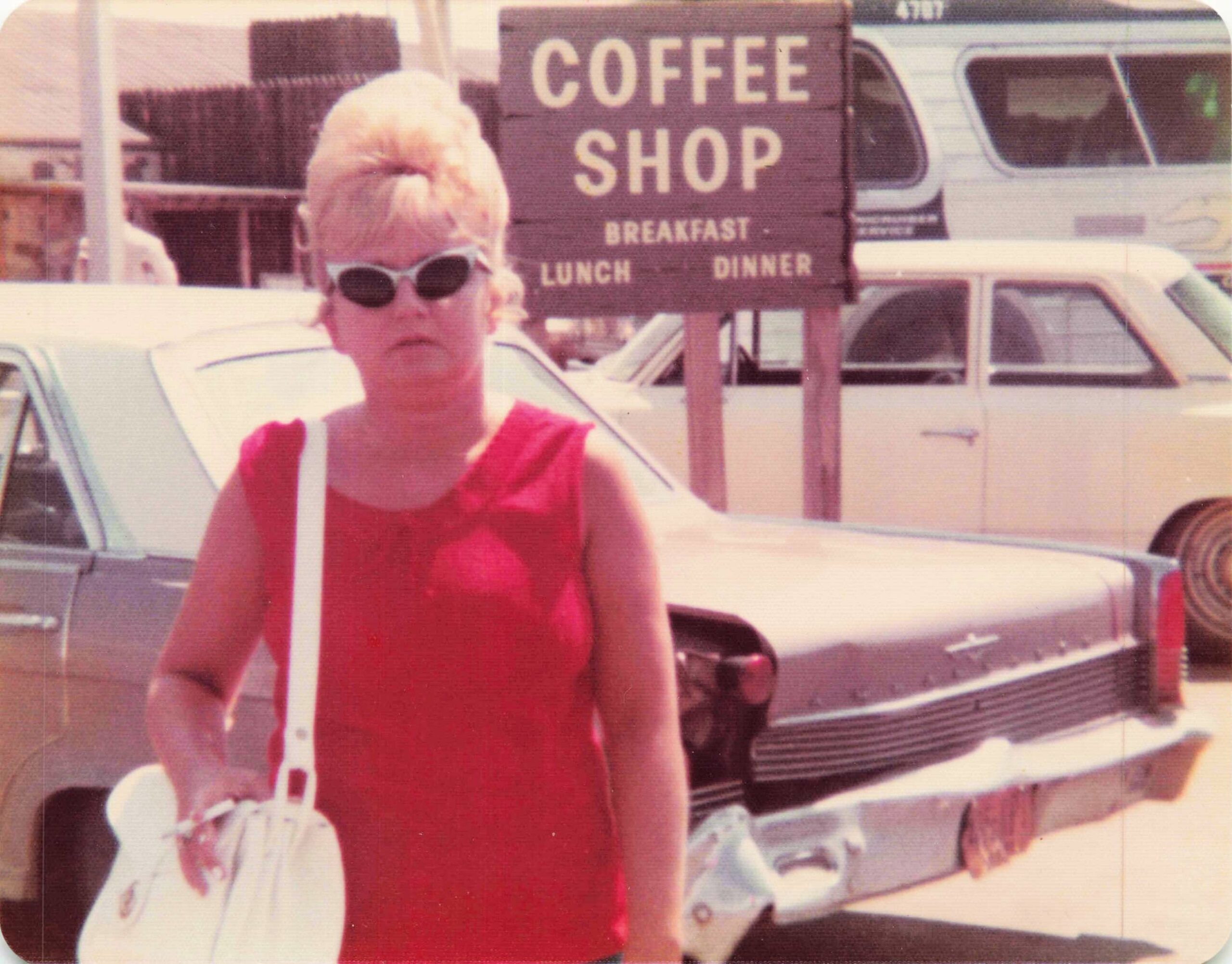 Person in a red sleeveless dress and sunglasses standing in front of a coffee shop sign reading 'COFFEE SHOP' with a vintage van in the background.