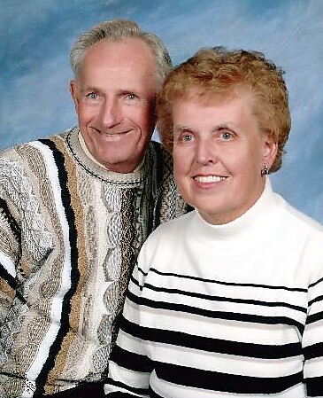 Smiling older couple posing together for a studio portrait, man in a patterned sweater and woman in a white‑black striped sweater, blue backdrop behind them.