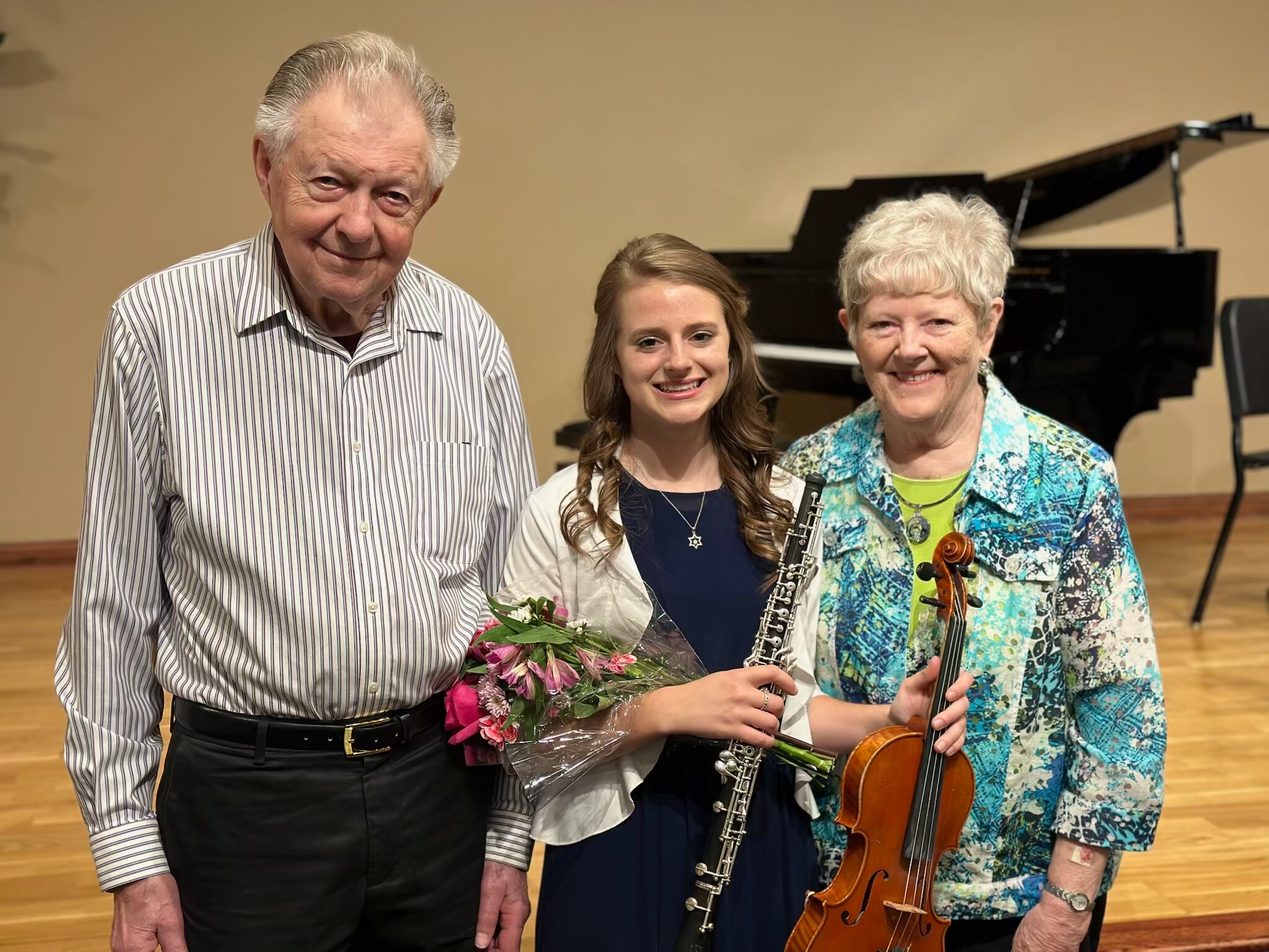 Three musicians posing together after a recital: an older man in a striped shirt, a young woman with a bouquet and a clarinet, and an older woman in a colorful top holding a violin; a piano in the background.