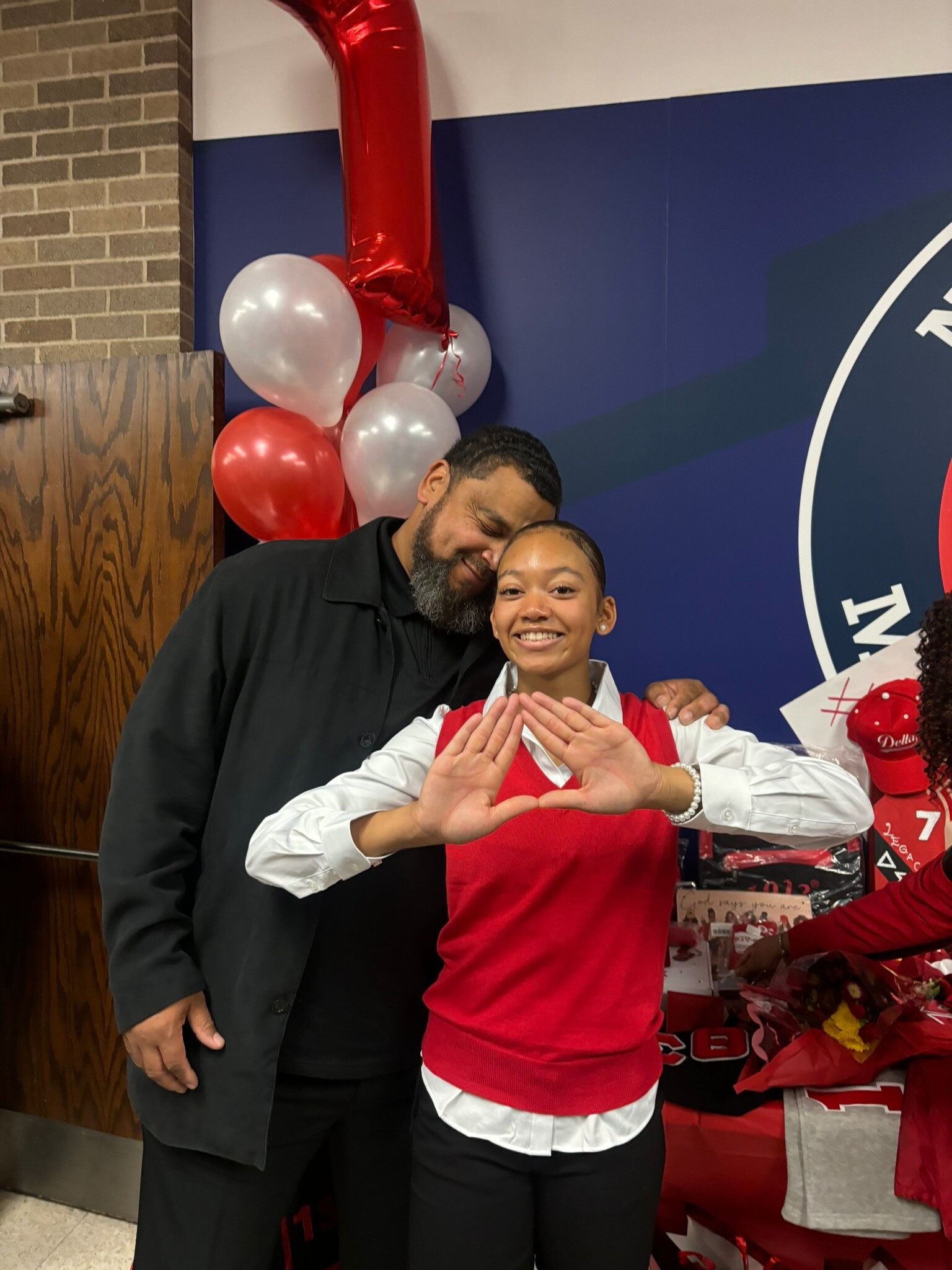 Smiling couple posing indoors with red and white balloons in the background; woman in a red sweater and white shirt, man in a dark coat, both making a triangle with their hands.