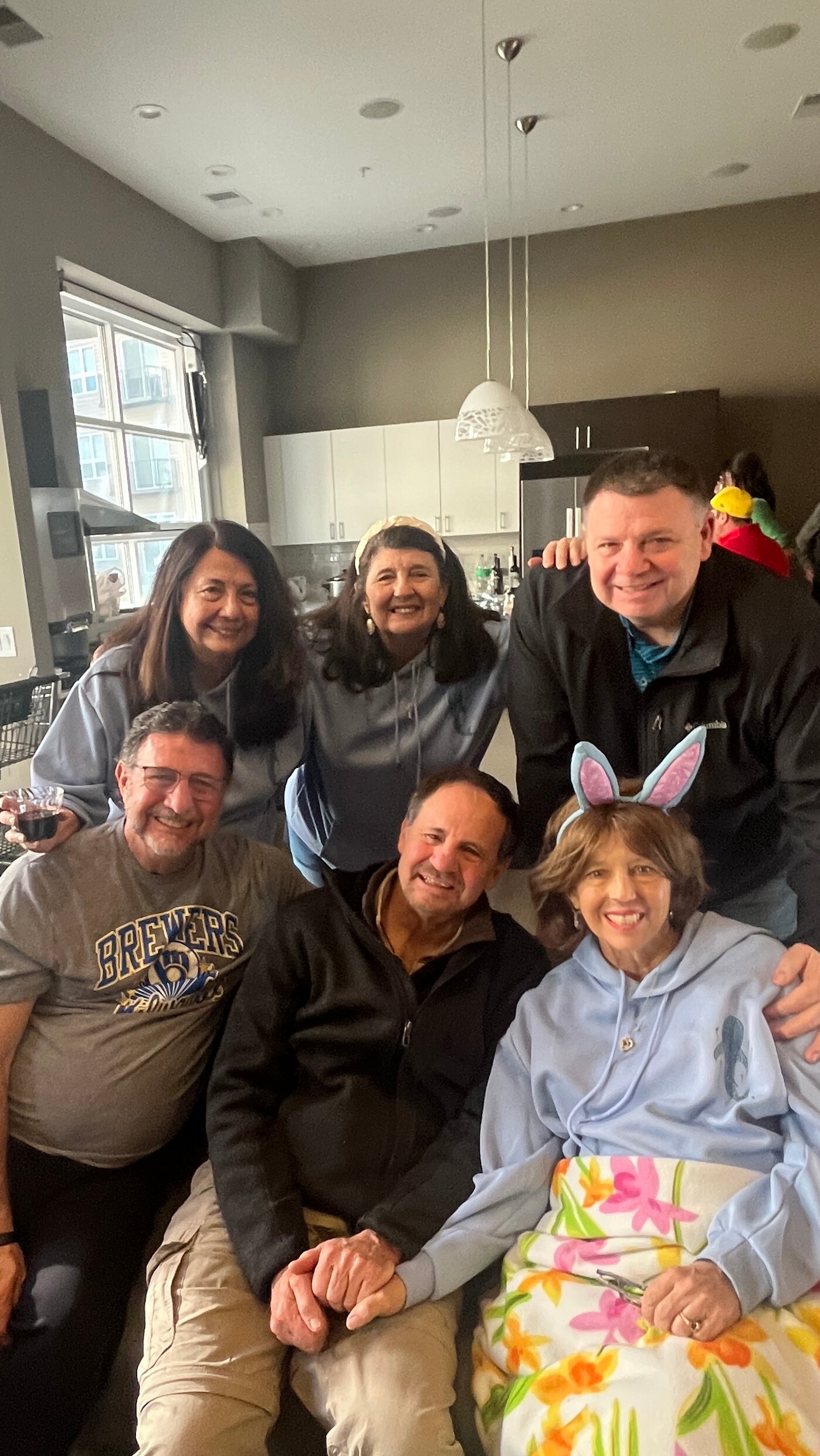 Group of adults posing and smiling for a casual kitchen gathering; one person wears bunny ears, others in relaxed clothing.