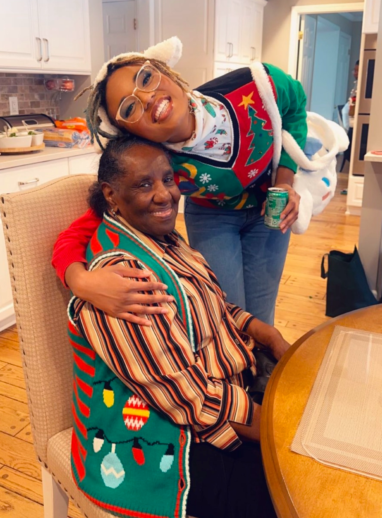 Two women share a hug in a bright kitchen during Christmas; one seated in a striped shirt, the other in a festive Christmas sweater with glasses, holding a soda can.