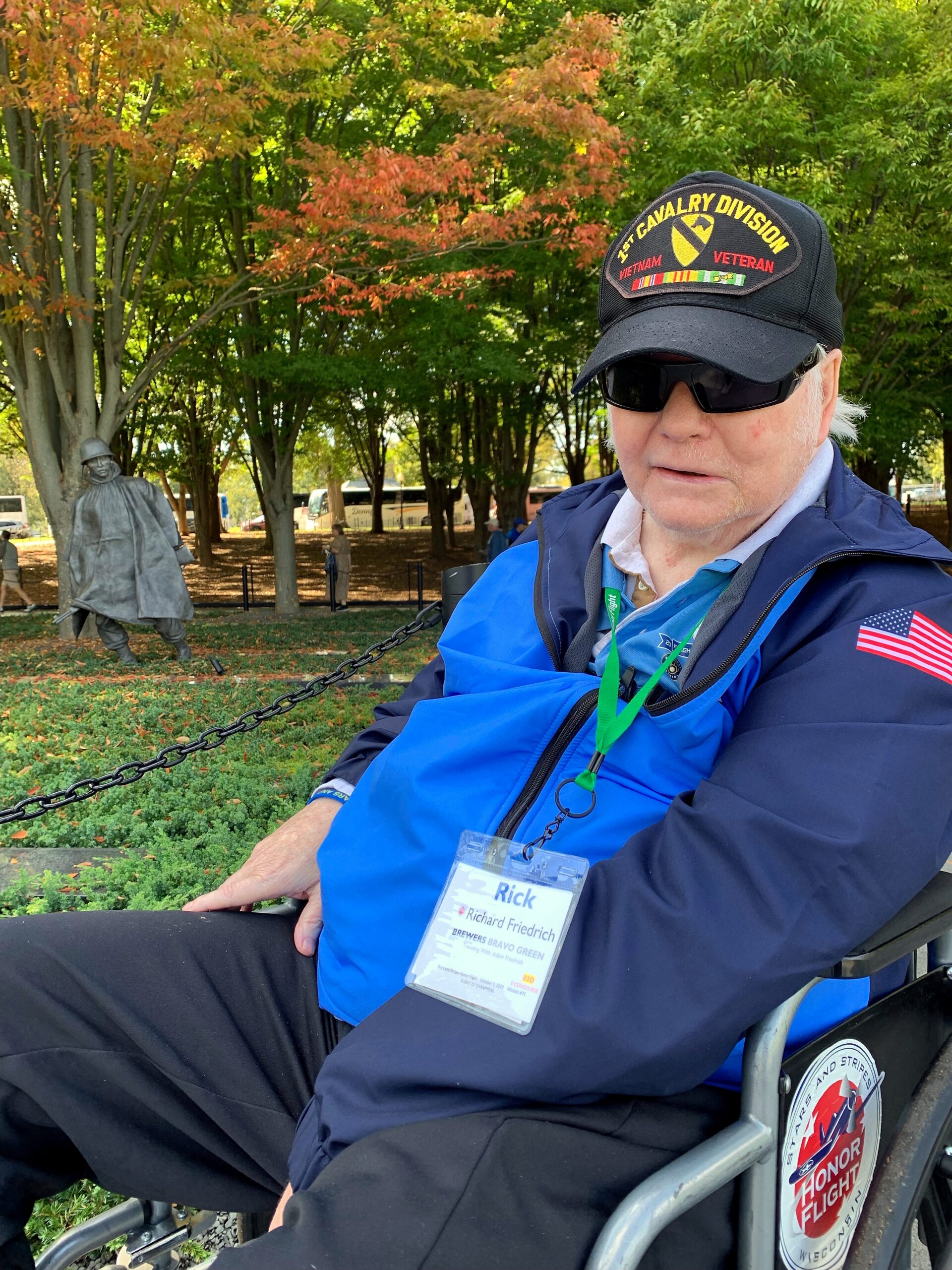 Older man in a blue jacket and sunglasses sits in a wheelchair in a park, wearing a name badge and a cap with veterans insignia; fall trees in the background.