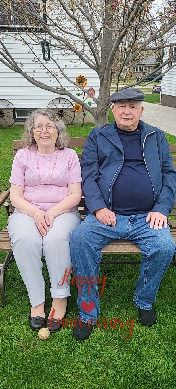 Elderly couple sits on a wooden bench in a yard, smiling; a tree and decorative sunflowers behind them, with pink script reading 'Happy Anniversary' overlaid on the grass.