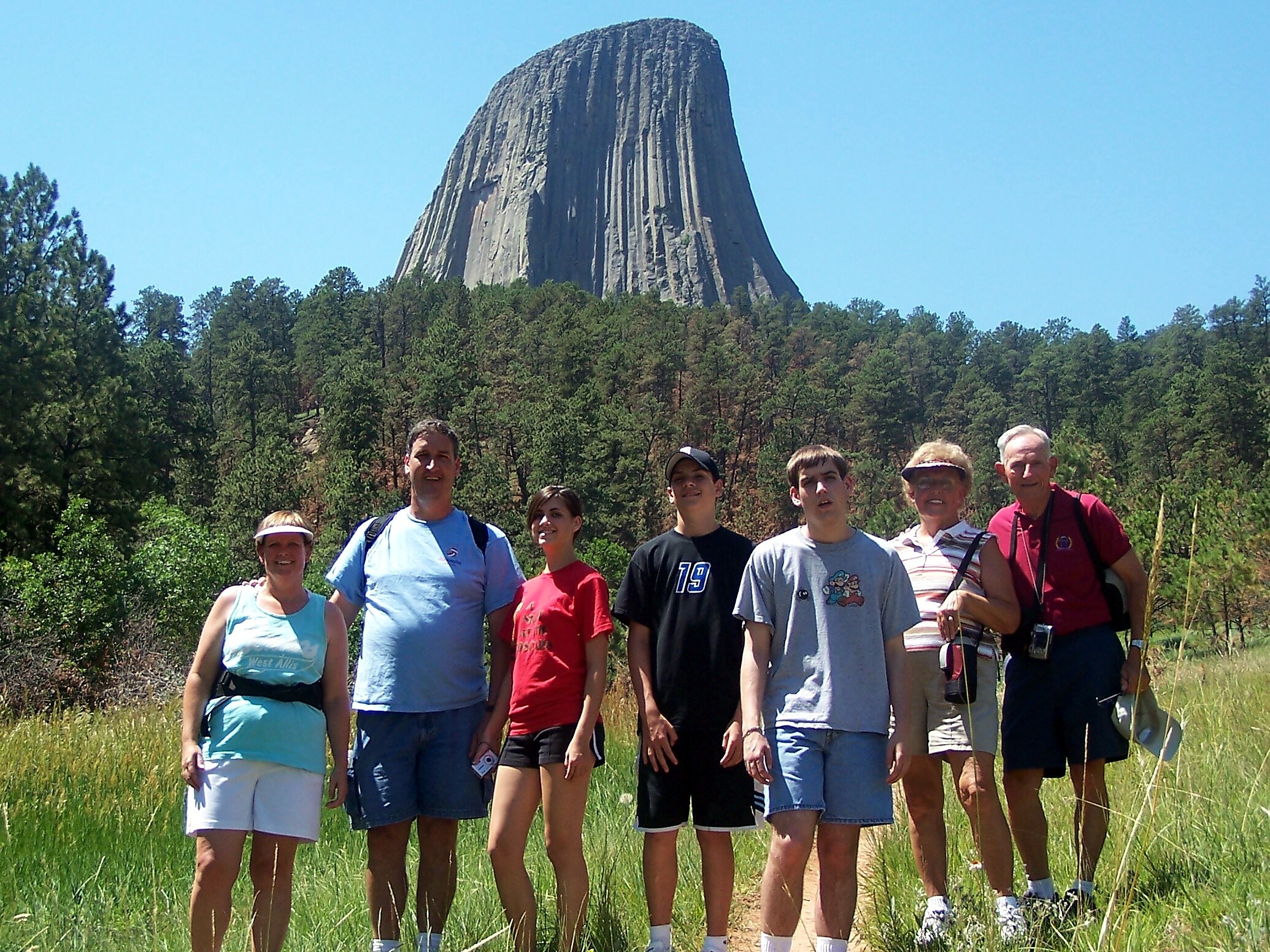 Group of people posing outdoors in a sunny field with a tall rock formation and trees behind them.