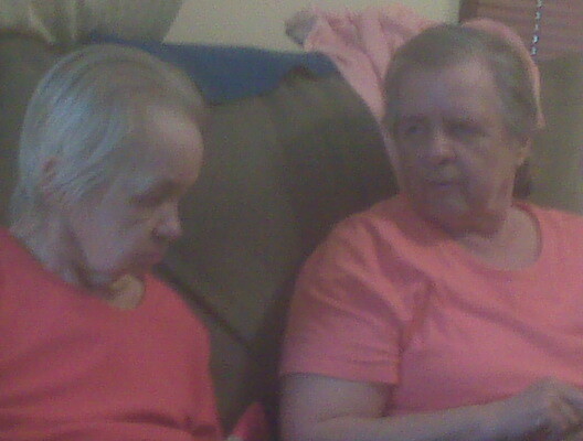 Two elderly women wearing pink shirts sit on a sofa and talk to each other.