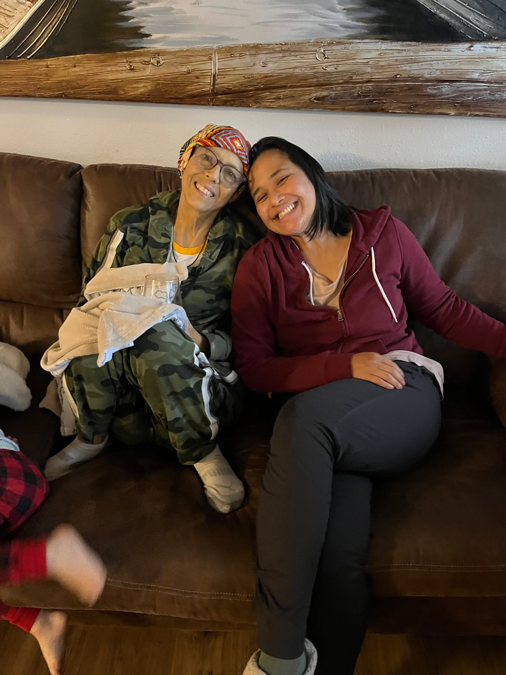 Two women sit close on a brown couch, smiling at the camera: one in camouflage clothing with a colorful headscarf, holding a blanket; the other in a burgundy hoodie.