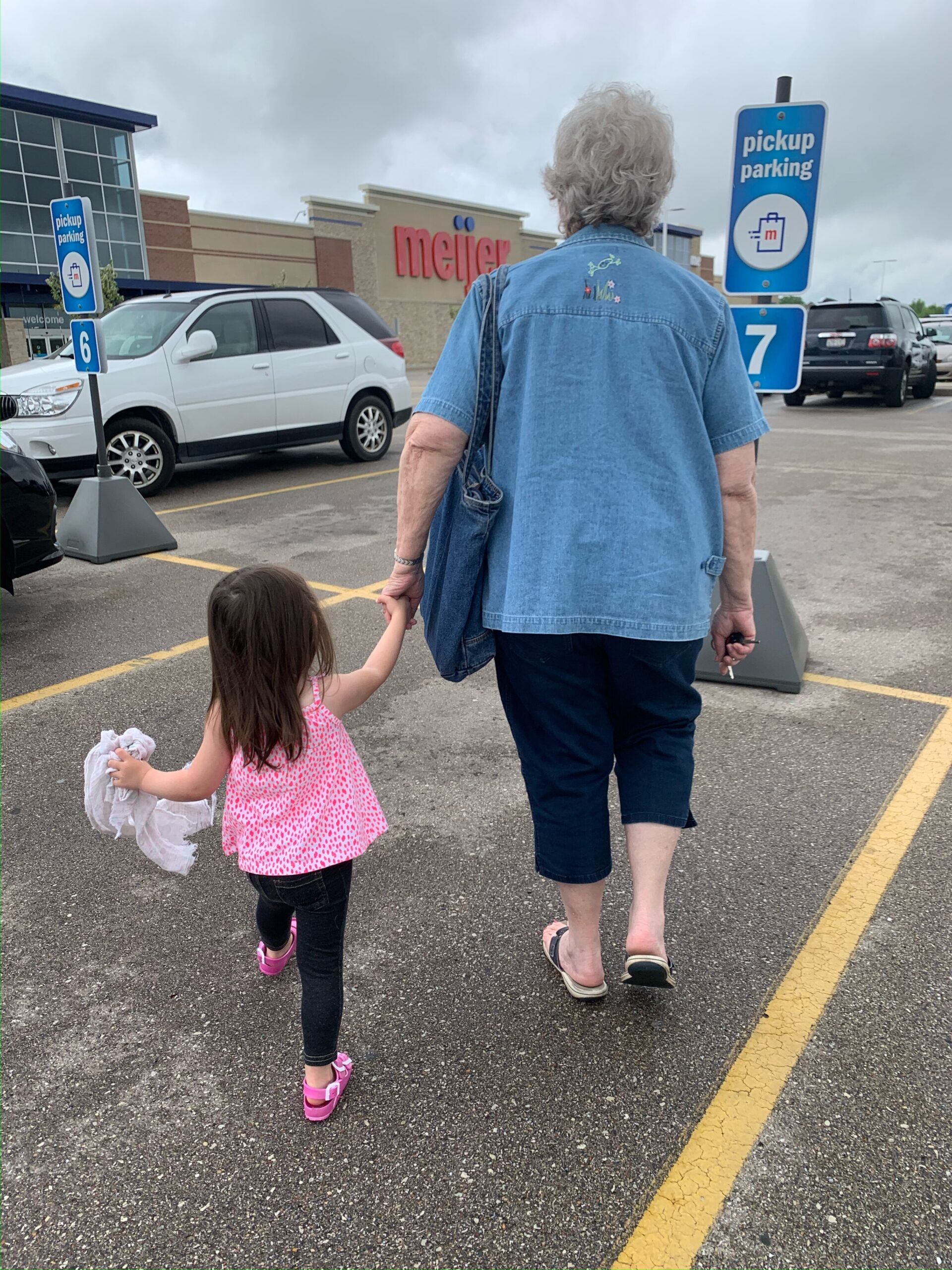 Back view of an older woman with short gray hair in a denim shirt, holding a child’s hand as they walk through a Meijer parking lot.