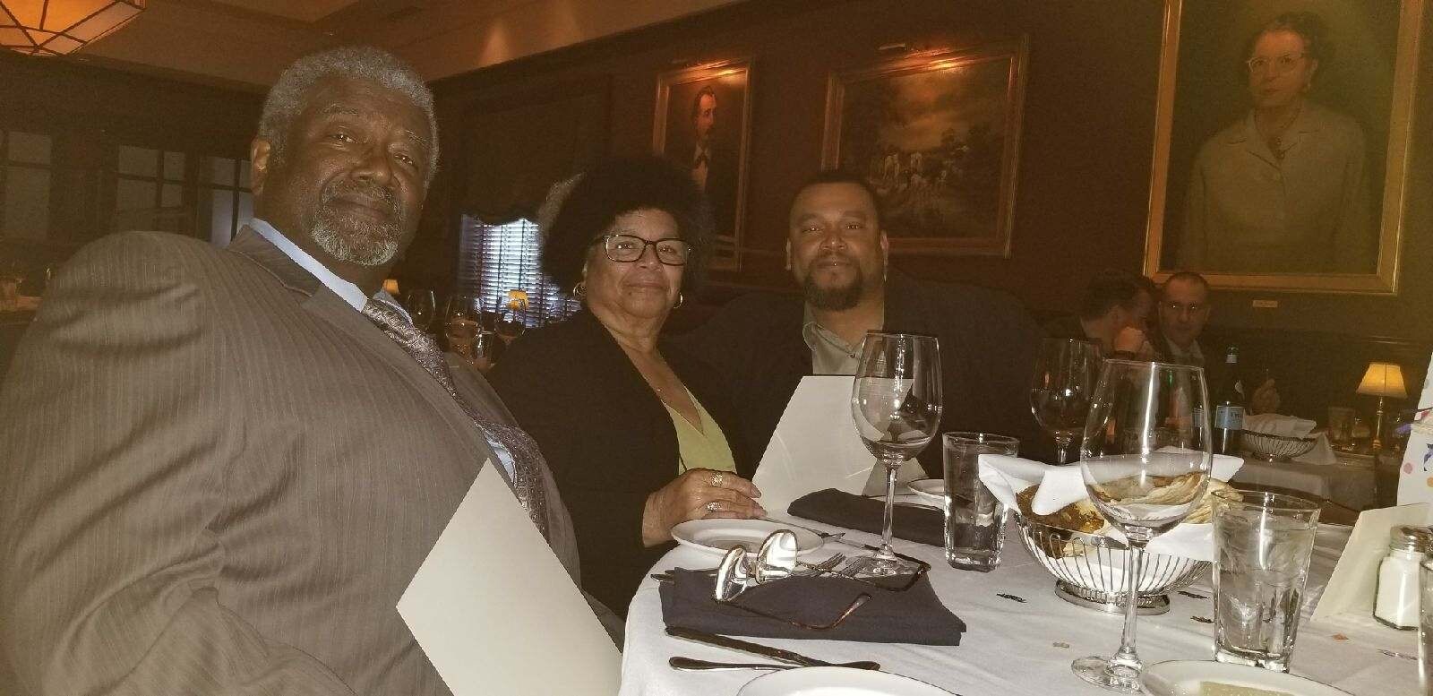 Three diners pose for a photo at a formal restaurant table with wine glasses and a bread basket.