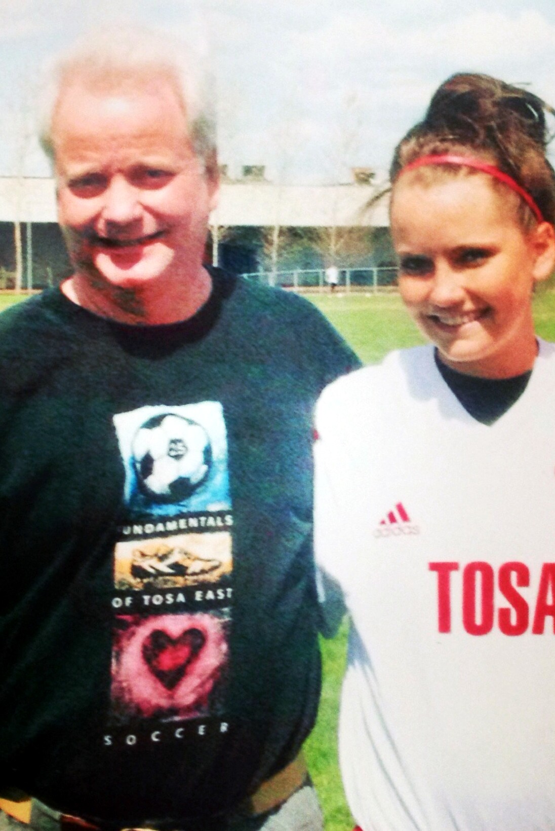 Two people smiling for a photo on a grassy soccer field, a man in a black T-shirt with a soccer-themed graphic and a young woman in a white jersey beside him.