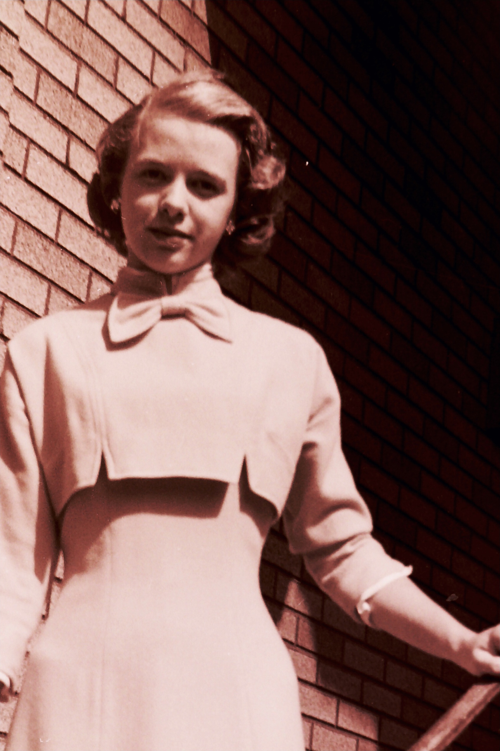 Young woman wearing a light-colored coat with a large collar, standing beside a brick wall.