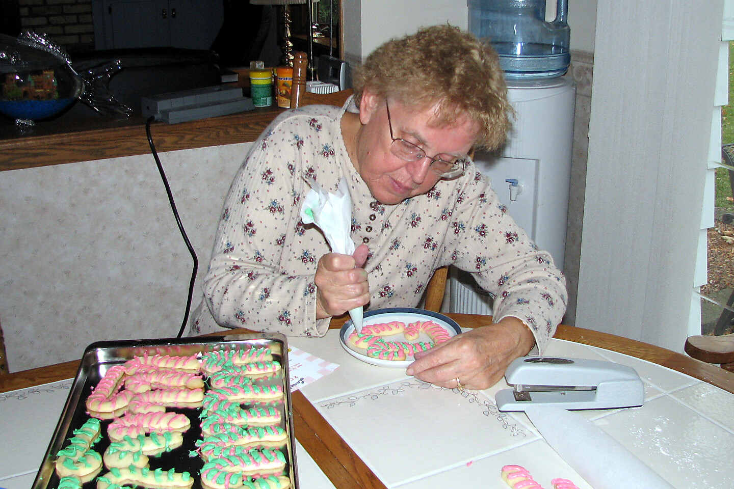Senior woman in floral pajamas pipes icing onto cookies at a kitchen table, focused on decorating.