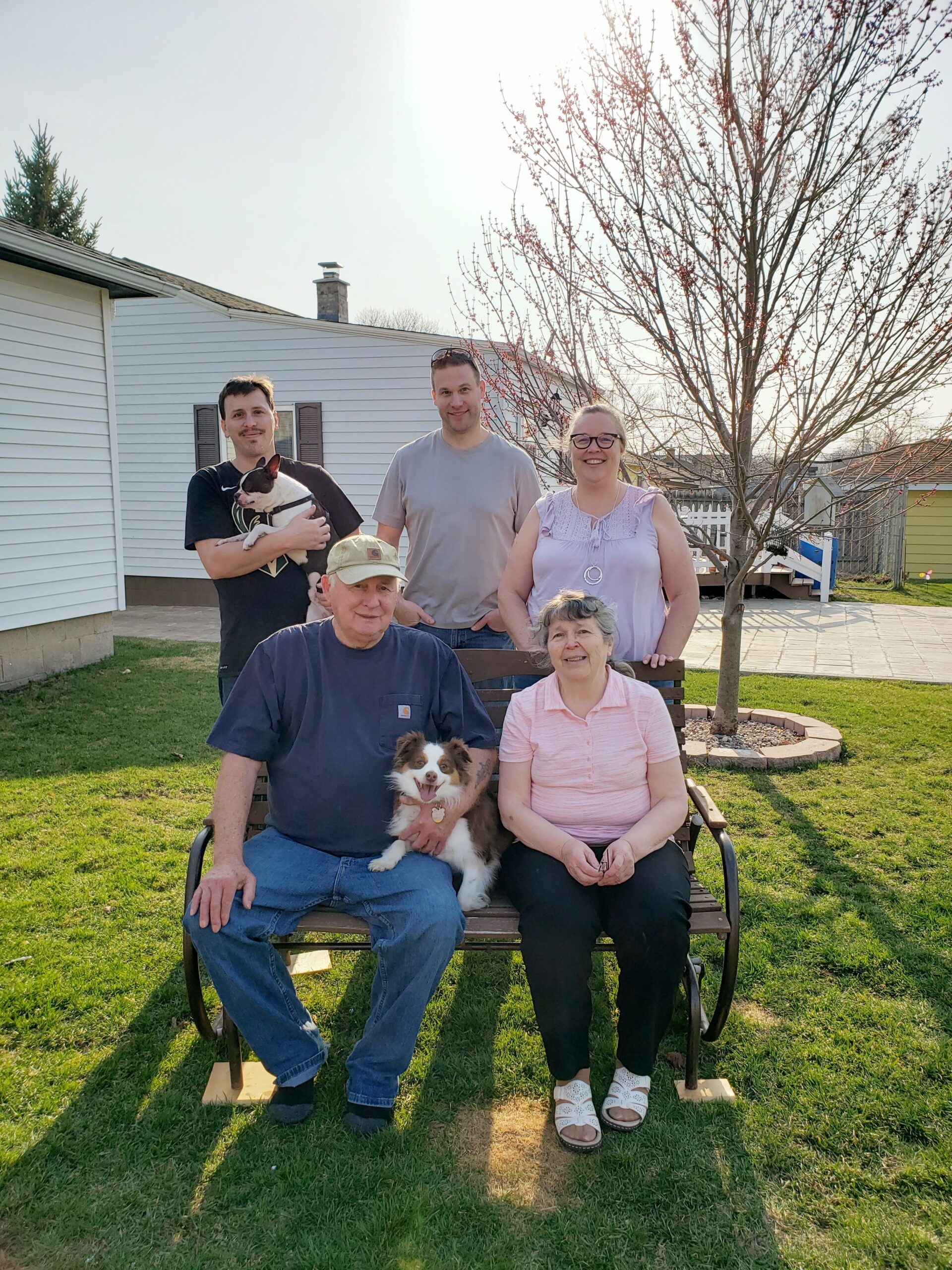 Five people (three adults standing, two seniors seated) pose in a sunny backyard with a small fluffy dog on the man’s lap, a tree and house in the background.