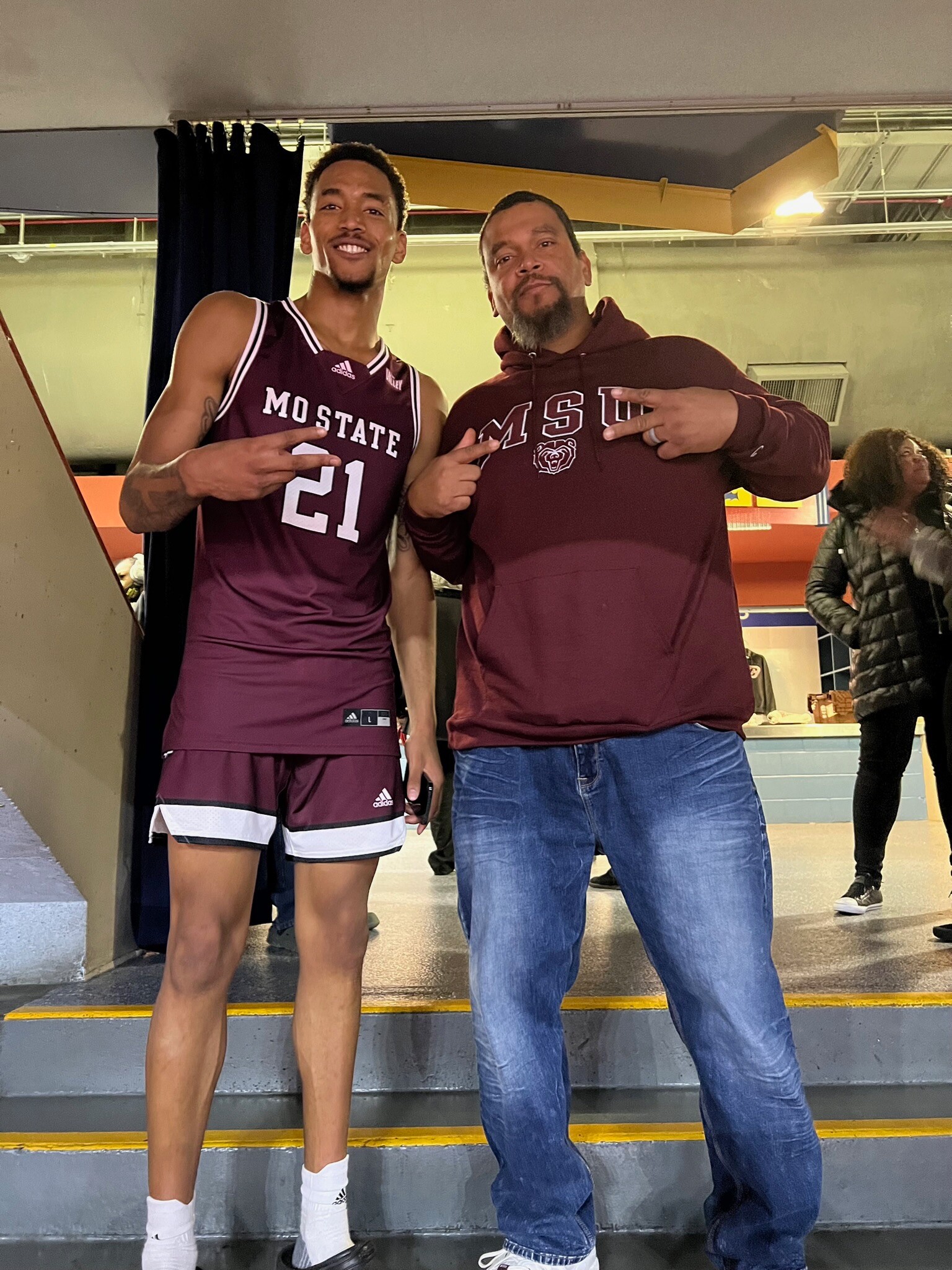 Two men pose on indoor stairs wearing MSU sports apparel, flashing peace signs at the camera