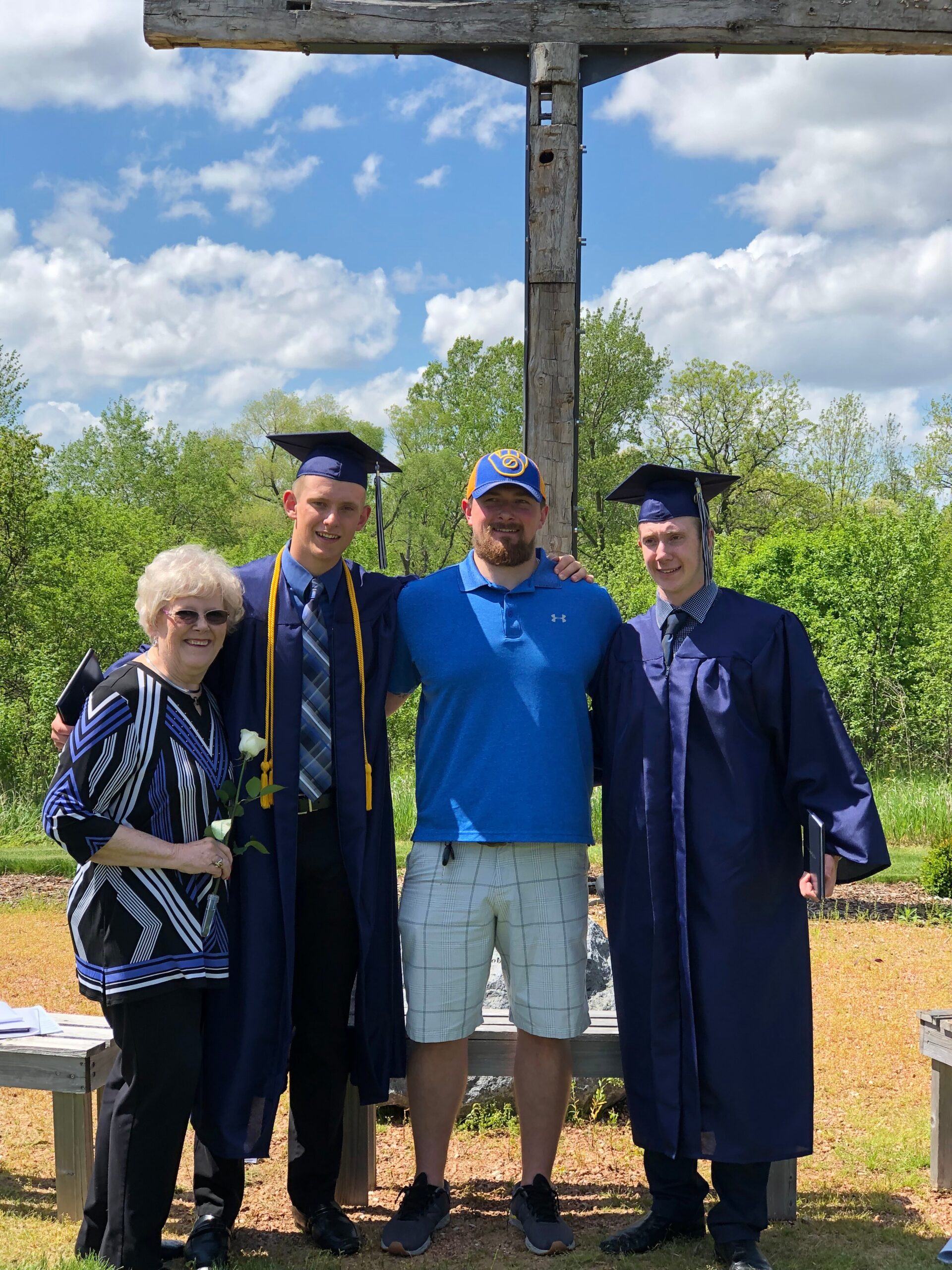 Four people pose together outdoors under a wooden structure: an elderly woman in pattern top and two graduates in navy gowns with mortarboards, plus a man in a blue polo shirt.