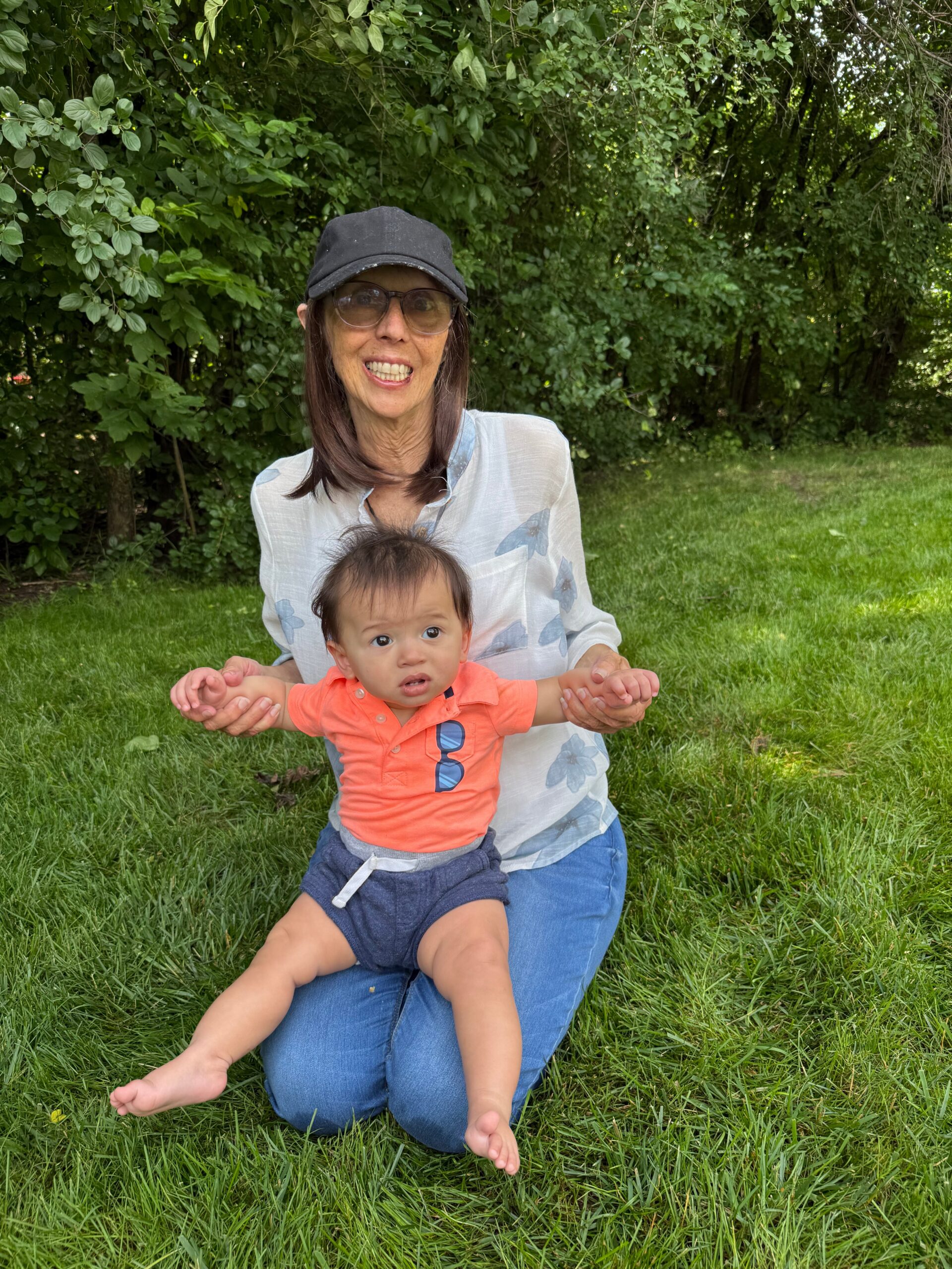 Woman wearing a black cap and glasses sits on grass, smiling, while holding a baby in an orange shirt outdoors near green bushes and trees.