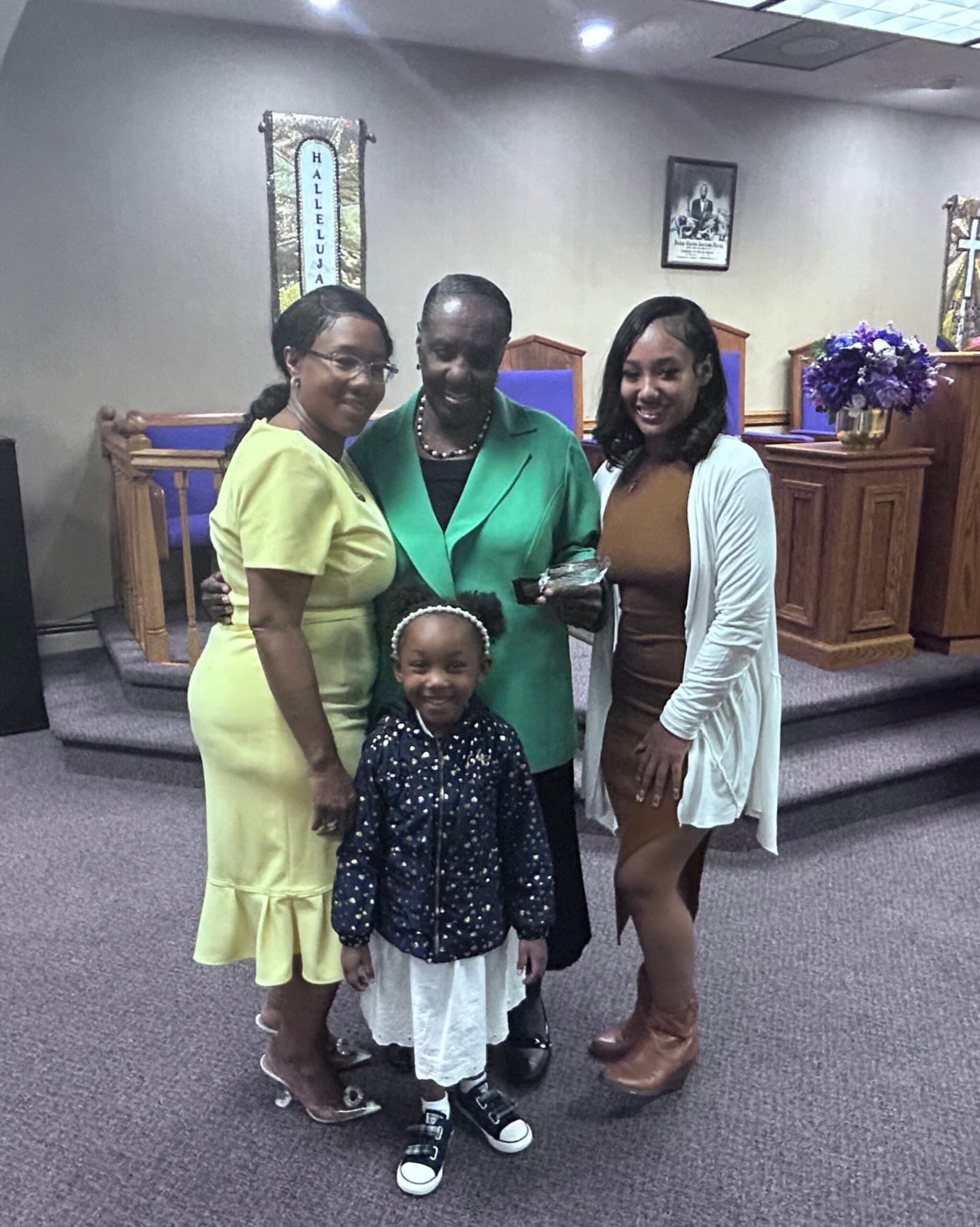 Four people posing and smiling in a church sanctuary, with a young girl in front wearing a navy polka-dot jacket and white skirt. Women stand behind her in bright outfits (yellow dress, green jacket, and brown dress with a white cardigan).