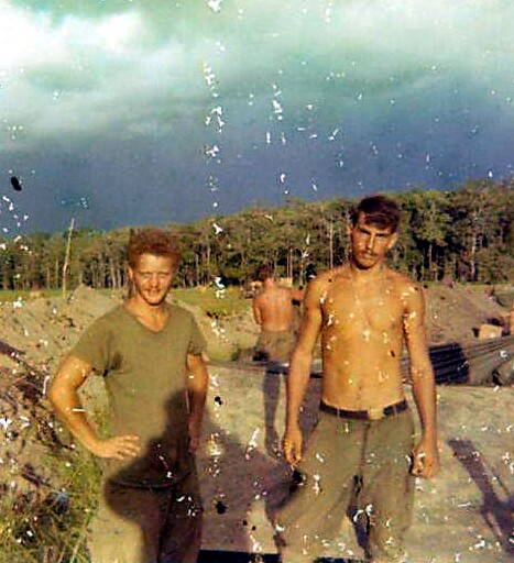 Two young men pose on a sandy beach with a treeline in the background; one wears a green t-shirt, the other is shirtless wearing green pants.