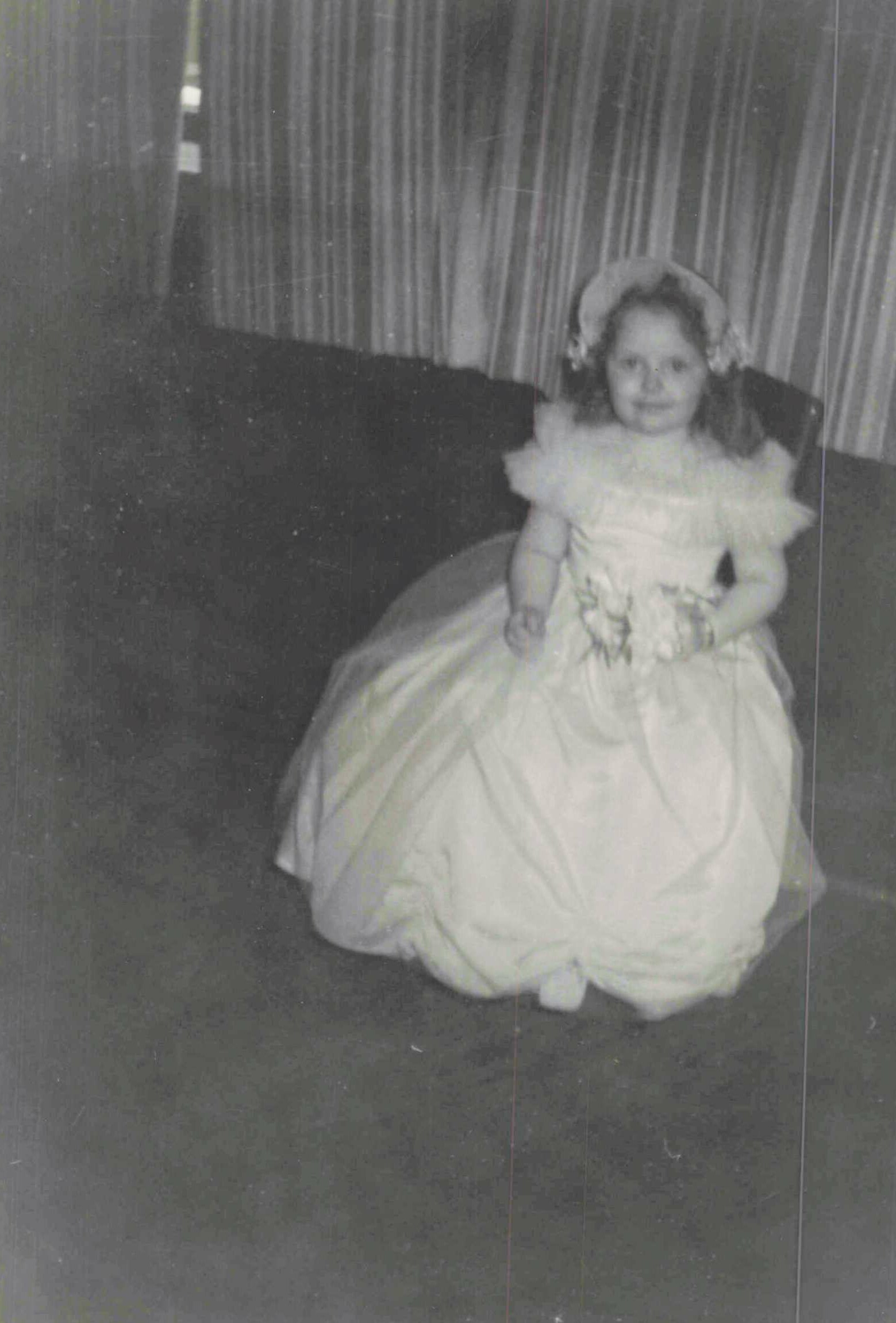 Young girl in a white formal gown with a fluffy headpiece, seated indoors holding a bouquet.