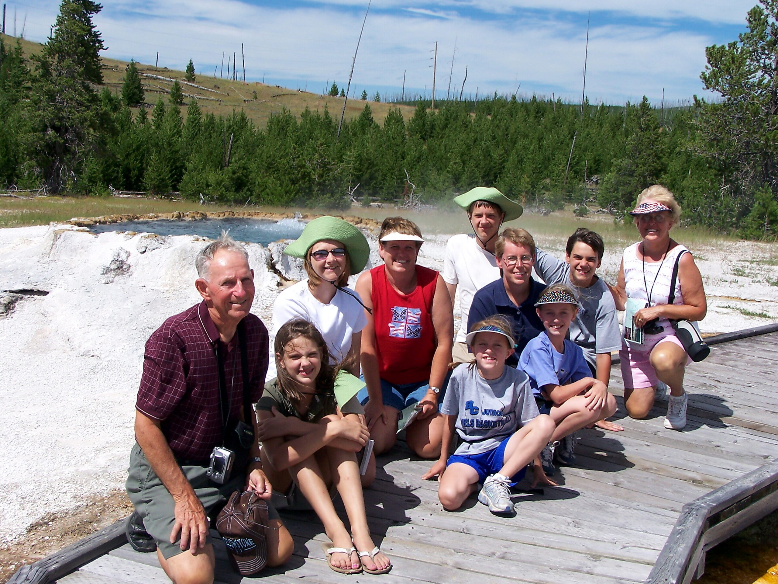 Group photo of adults and kids posing on a wooden boardwalk beside a white mineral terrace and steam, with pine forest in the background.