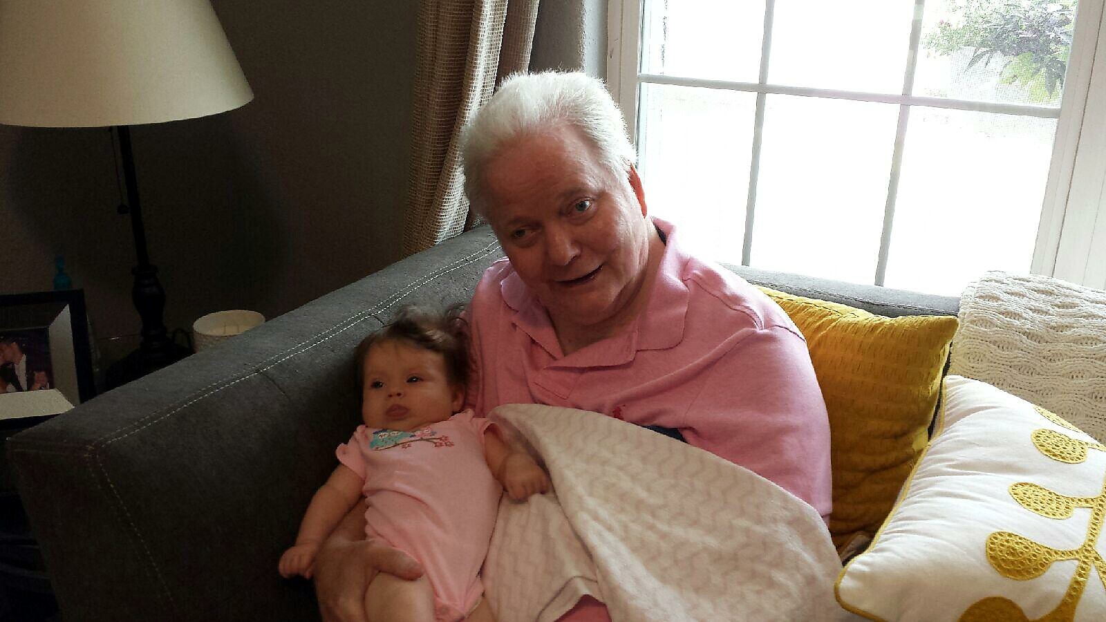 Older woman in a pink polo shirt sits on a gray sofa, cradling a baby in a pink onesie with a white blanket nearby by a sunlit window.] | note: concise informative alt text for a warm familial moment, showing relationship and setting but not overly descriptive.