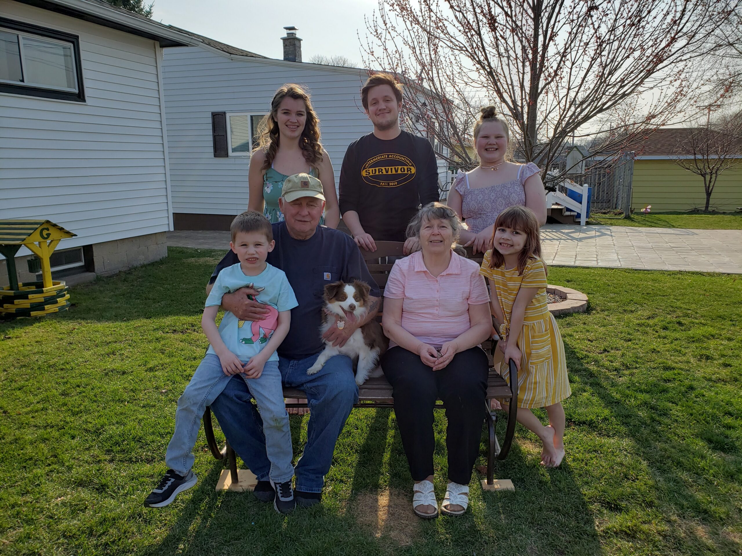 Family group of adults and children posing on a bench in a sunny backyard, with a small dog in front.