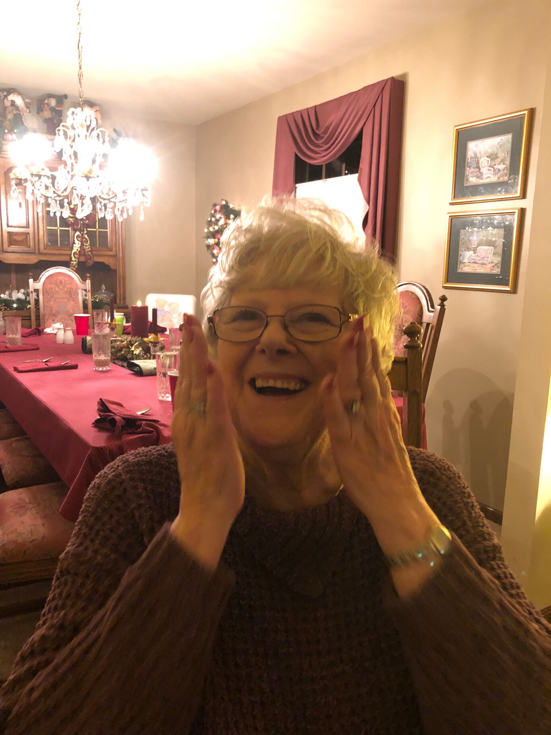 Older woman with glasses smiling and cupping her face at a festive Christmas dinner table.