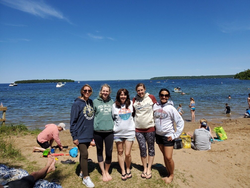 Five smiling women stand arm in arm on a sunny sandy beach by a calm lake, with boats and people playing in the water in the background.
