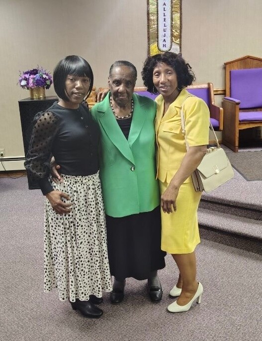Three women stand together in a church interior, arm in arm, smiling at the camera. The center woman wears a green blazer with black skirt, flanked by a woman in a black polka-dot dress and a woman in a yellow dress with a beige bag.