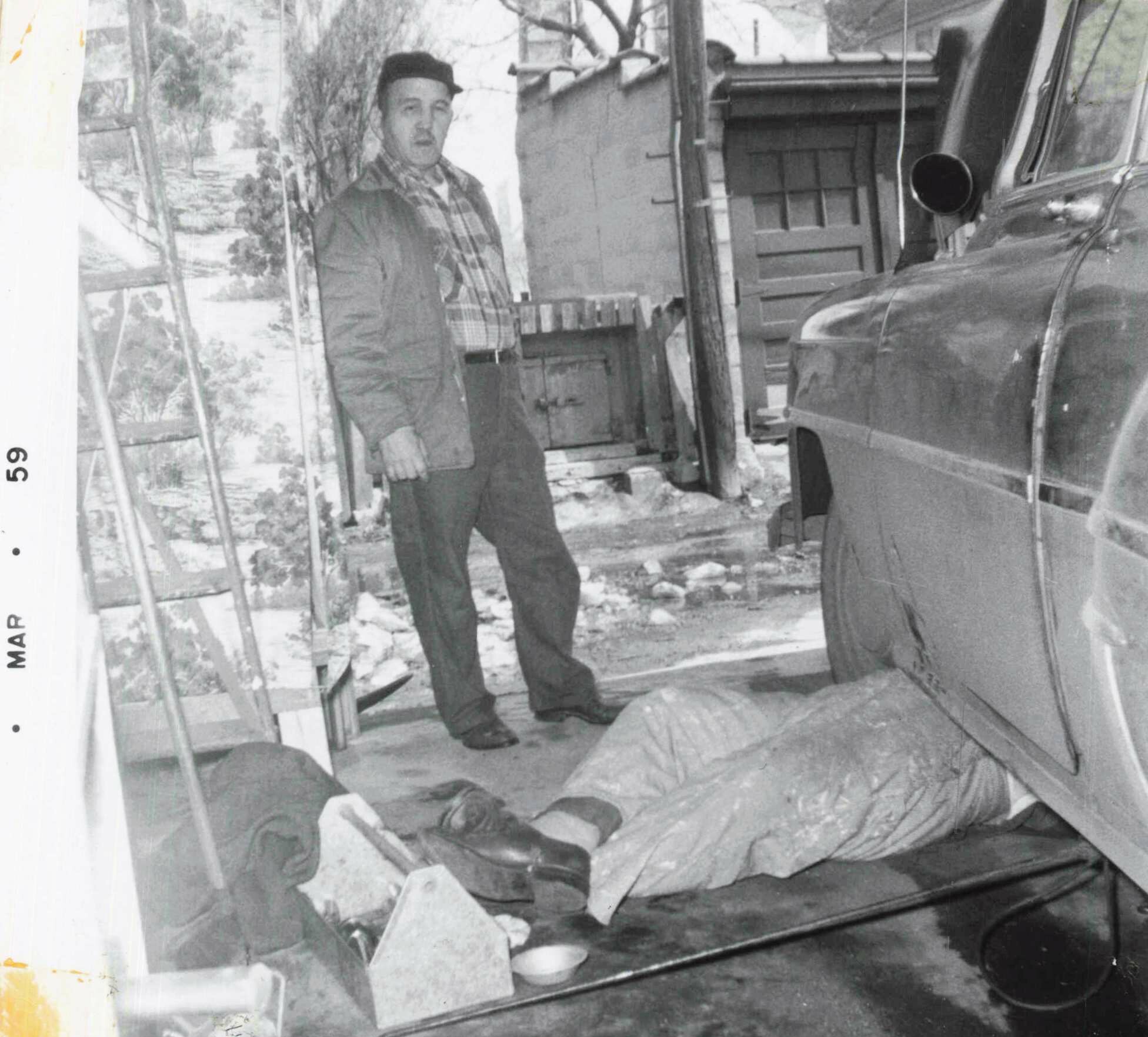 Black-and-white photo of a man in a hat and scarf standing beside a large truck in a rubble-filled outdoor scene, looking at the camera.